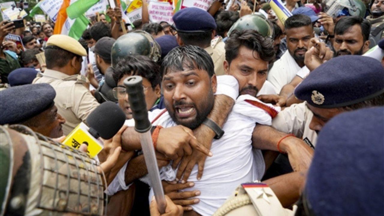 Police personnel attempting to restrain and evict a male protester amid a large gathering during a student protest in Patna. Several uniformed police officers are seen holding the protester, while other protesters and police personnel crowd the area in the background. Protesters can be seen carrying flags and placards during the march.